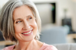 A smiling senior woman with gray hair looks off to the side, showing confidence and happiness, possibly after receiving dental implants or restorative dental care.