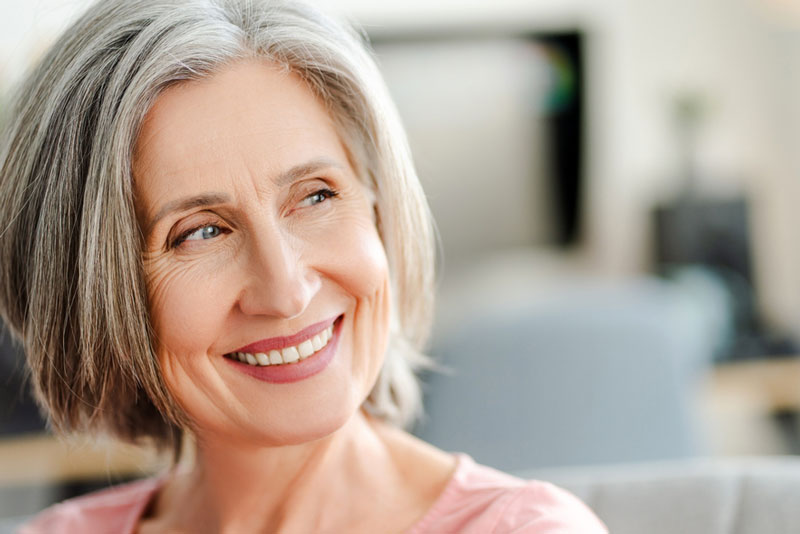 A smiling senior woman with gray hair looks off to the side, showing confidence and happiness, possibly after receiving dental implants or restorative dental care.